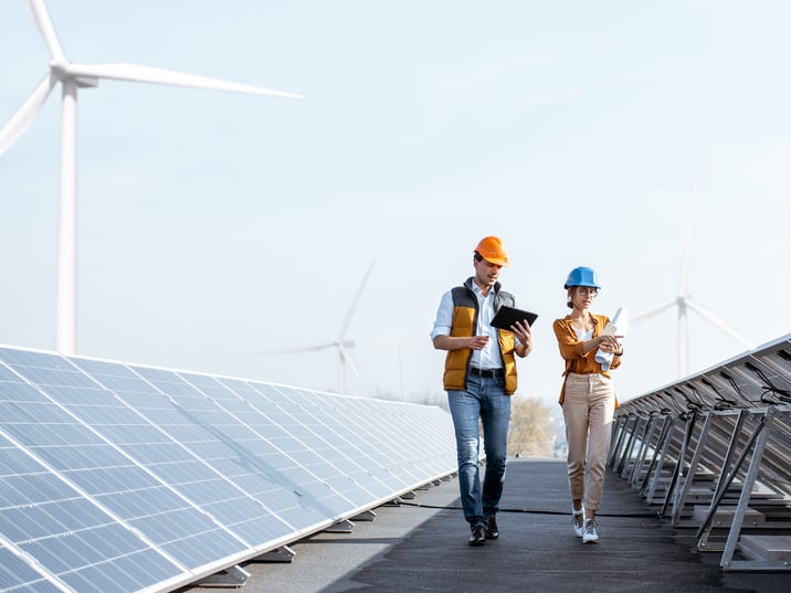 Two workers walking alongside solar panels with wind turbines in the background